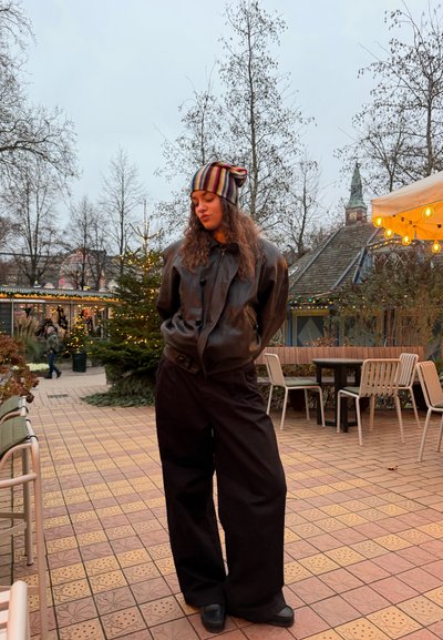 Joven mujer con chaqueta de cuero y gorro de punto colorido, de pie con las manos en los bolsillos en un patio al aire libre con baldosas, árboles y decoraciones iluminadas.
