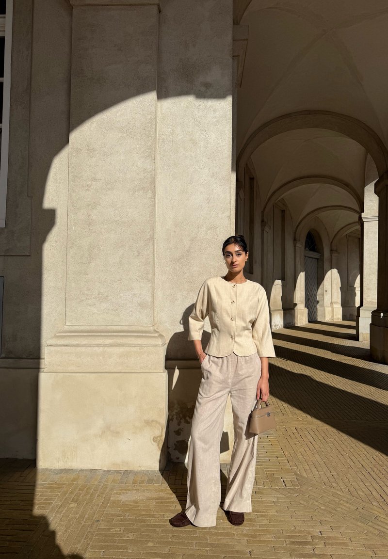 A woman in a beige top and trousers is holding a small handbag and stands against a sunlit pillar in an arched corridor, where patterned shadows play across the surfaces.