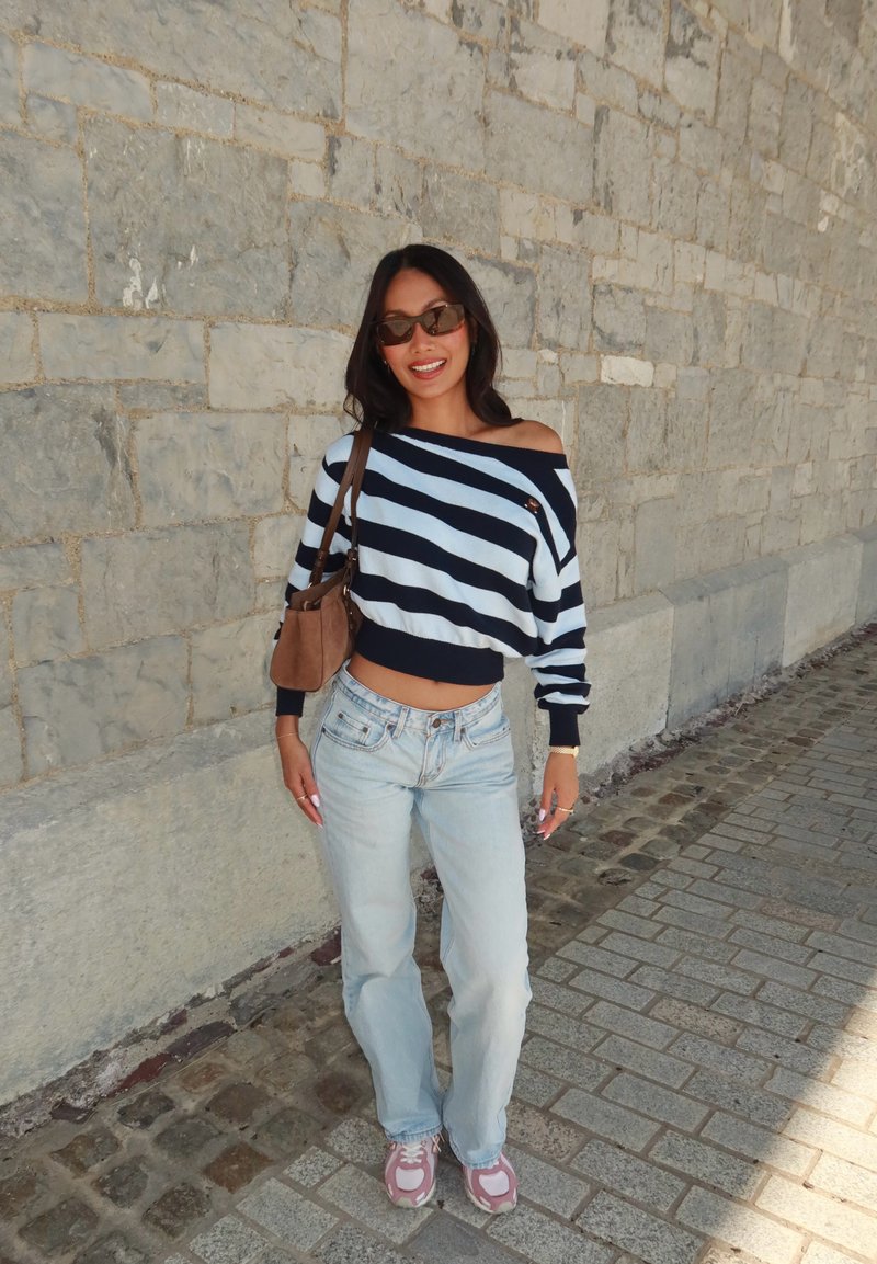 Donna con capelli lunghi e scuri che indossa occhiali da sole, maglione a righe con spalle scoperte, jeans chiari, sneakers rosa e borsa a tracolla marrone, in piedi vicino a un muro di pietra.