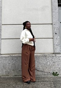 Felpa bianca con zip, pantaloni larghi marroni, orecchini a cerchio dorati e bracciale. In piedi contro un muro grigio testurizzato. Tiene degli occhiali da sole.