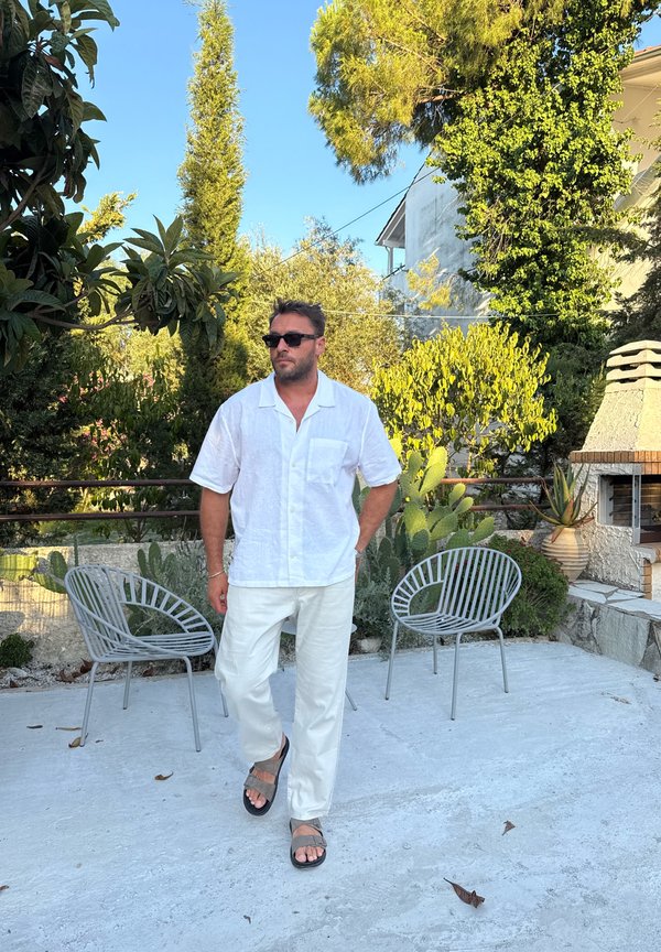 Hombre con camisa y pantalones blancos que lleva gafas de sol y sandalias camina por un patio con dos sillas de metal y plantas verdes bajo la luz brillante del sol.