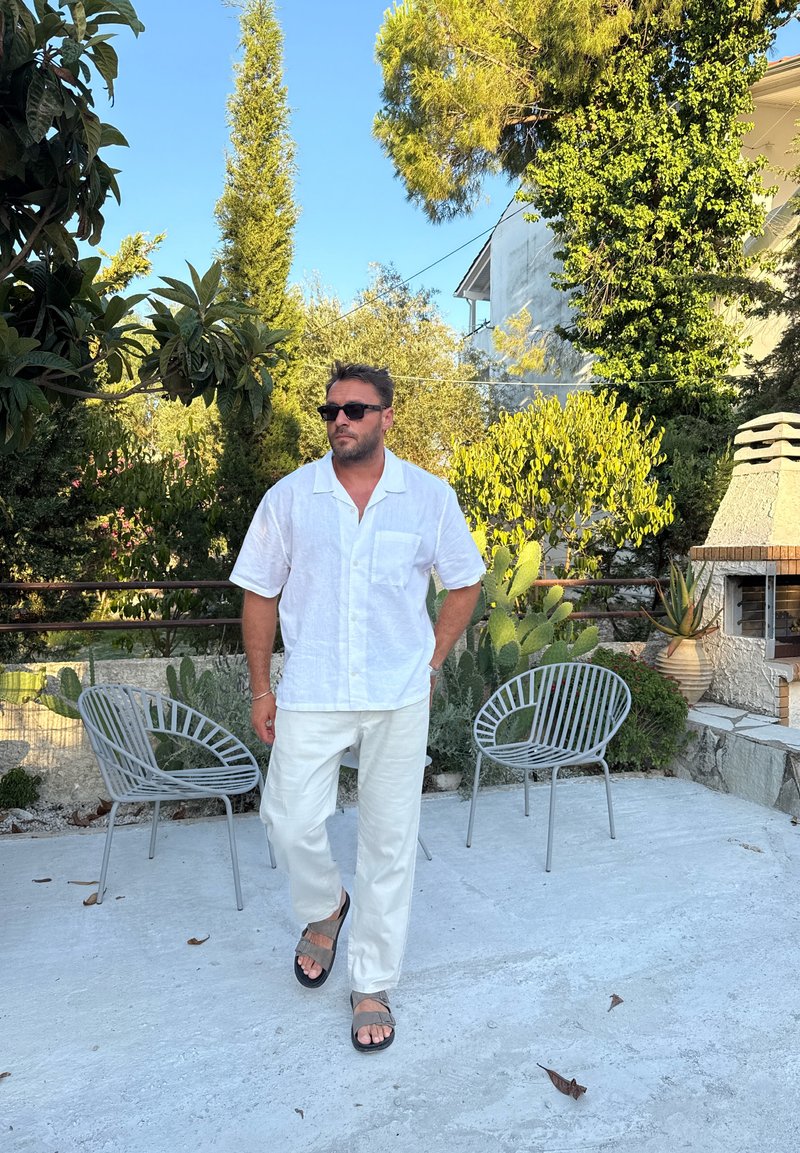 Man in white shirt and pants wearing sunglasses and sandals walks on patio with two metal chairs and green plants in bright sunlight.