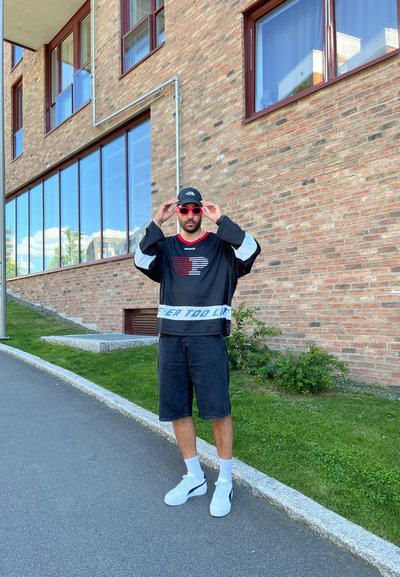 Camisa negra de manga larga con gráfico rojo, mangas blancas; pantalones cortos negros; zapatillas deportivas blancas; gafas de sol rojas; de pie en una acera cerca de un edificio de ladrillo.