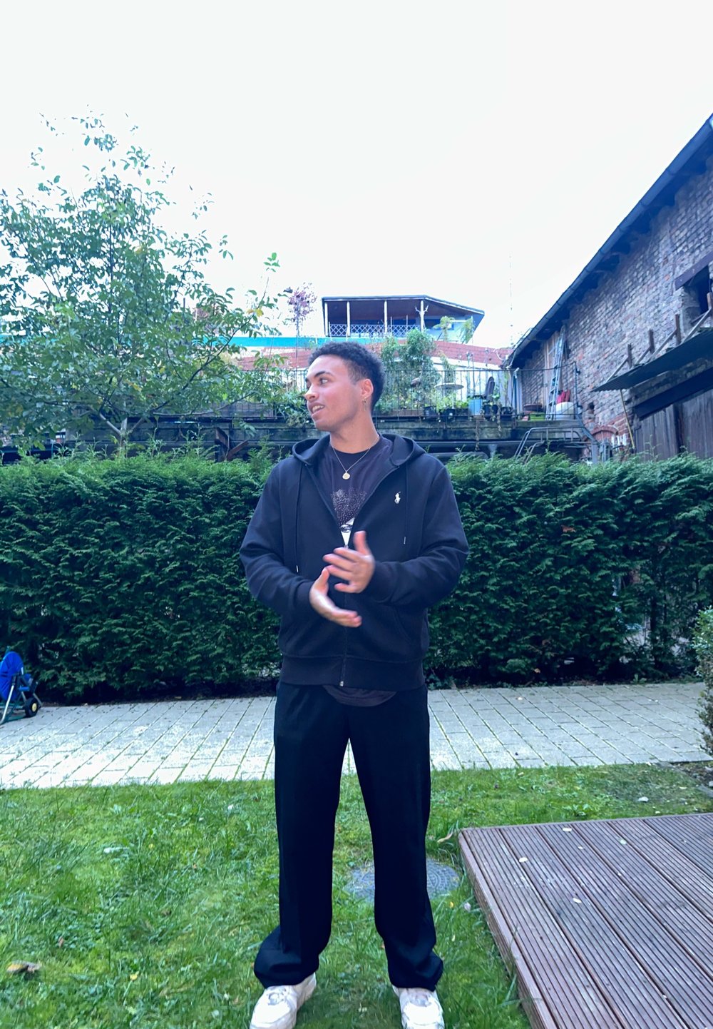 Melnā zipa kapuce virs grafiskās T-krekla, savienota ar melniem biksēm un baltām kedām, stāv dārzā ar zaļumiem un bruģakmeņiem.