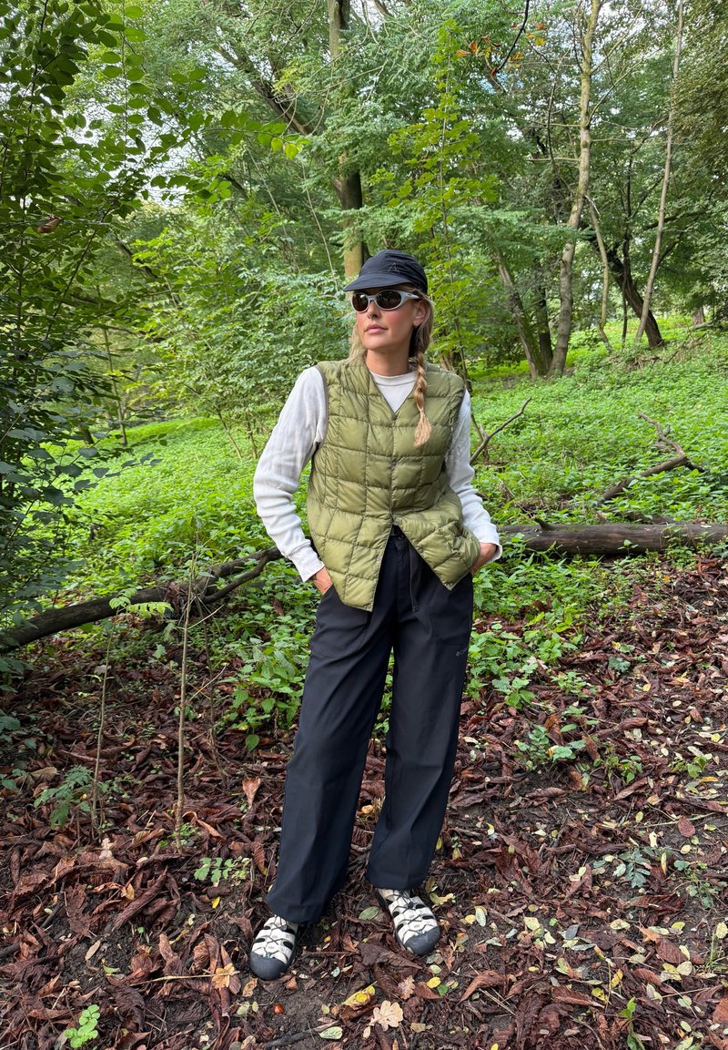 Mulher de óculos de sol, chapéu preto, colete acolchoado verde, camisa de manga longa branca, calças pretas e botas de caminhada, de pé numa área florestal com folhas.