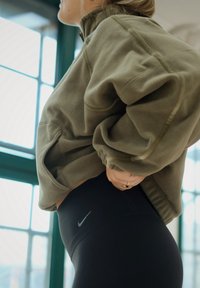 Veste en polaire vert olive avec un col montant, associée à des leggings noirs arborant un petit logo Nike. Texture lisse, coupe décontractée.