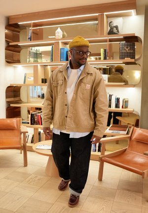 Khaki jacket with front pockets, layered over a white shirt, dark denim jeans, and brown shoes. An olive-green beanie completes the look.