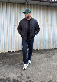 Fekete bombázó dzseki fekete ing fölött, kék farmernadrág és fehér tornacipő. Zöld sapka és tükröződő napszemüveg teszi teljessé a ruházatot.