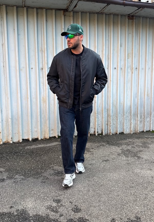 Jaqueta bomber preta sobre uma camisa preta, jeans azuis e ténis brancos. Boné verde e óculos de sol espelhados completam o visual.