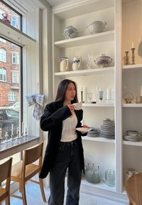 Une femme en blazer noir tient une tasse blanche et une assiette, debout près d'un présentoir de vaisselle en céramique et d'objets en verre sur des étagères.