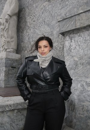 Mujer con cabello rizado oscuro que lleva una chaqueta de cuero negra y una bufanda gris claro, de pie contra una pared de piedra gris con una estatua tallada cercana.