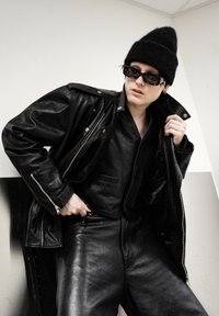 Veste en cuir noir avec fermetures éclair, superposée sur une chemise et un pantalon en cuir noir. Accessoirisée avec des lunettes de soleil foncées et un bonnet noir.