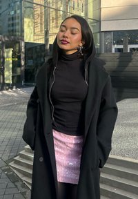 Langer schwarzer Mantel mit Kapuze über einem schwarzen, gerippten Rollkragenpullover, kombiniert mit einem schimmernden, rosa Paillettenrock. Im Hintergrund ist moderne Glasarchitektur zu sehen.