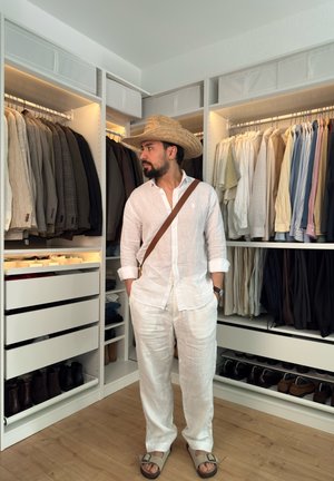Hombre con ropa de lino blanca y sombrero de paja de pie en un vestidor organizado con ropa colgada y zapatos sobre suelo de madera.