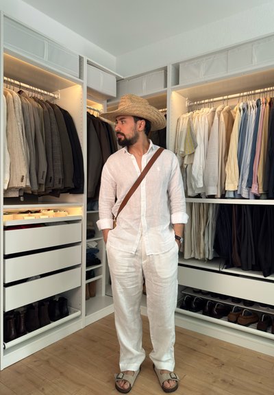 Hombre con ropa de lino blanca y sombrero de paja de pie en un vestidor organizado con ropa colgada y zapatos sobre suelo de madera.