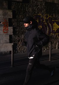 Veste imperméable noire avec col montant, leggings noirs avec logo Nike blanc, courant dans une rue de la ville, mur de pierre ombragé en arrière-plan.