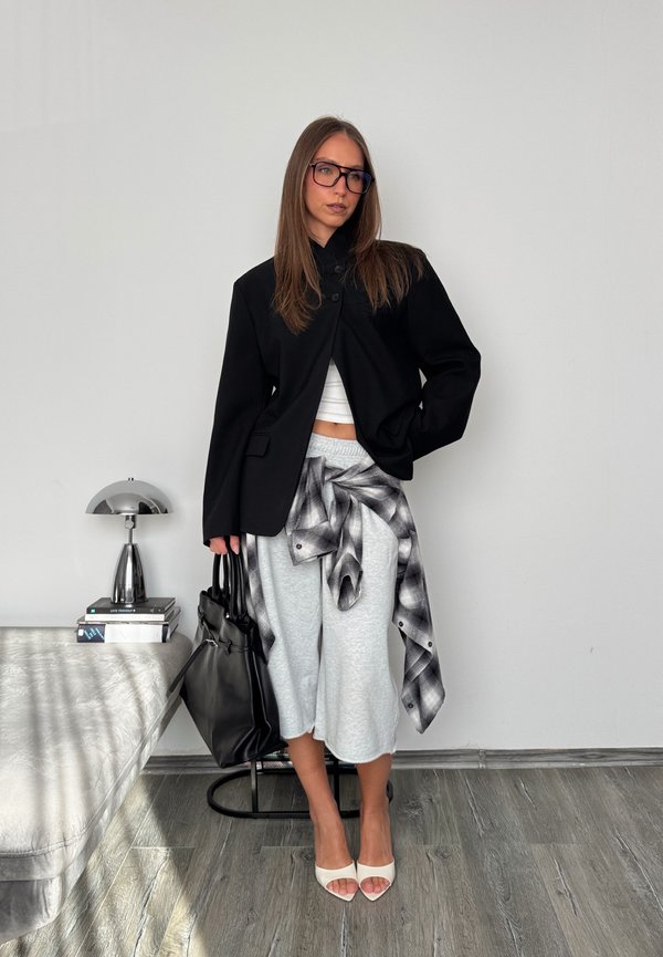 Femme portant un blazer noir, un haut blanc court, un short gris, une chemise à carreaux nouée à la taille, des talons blancs, tenant un sac à main noir, debout près d'une table avec lampe.