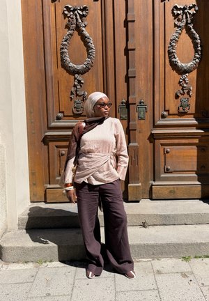 Mujer con camiseta beige, pantalones oscuros, pañuelo en la cabeza y gafas de sol, de pie en escalones de piedra frente a grandes puertas dobles de madera con tallas ornamentadas.