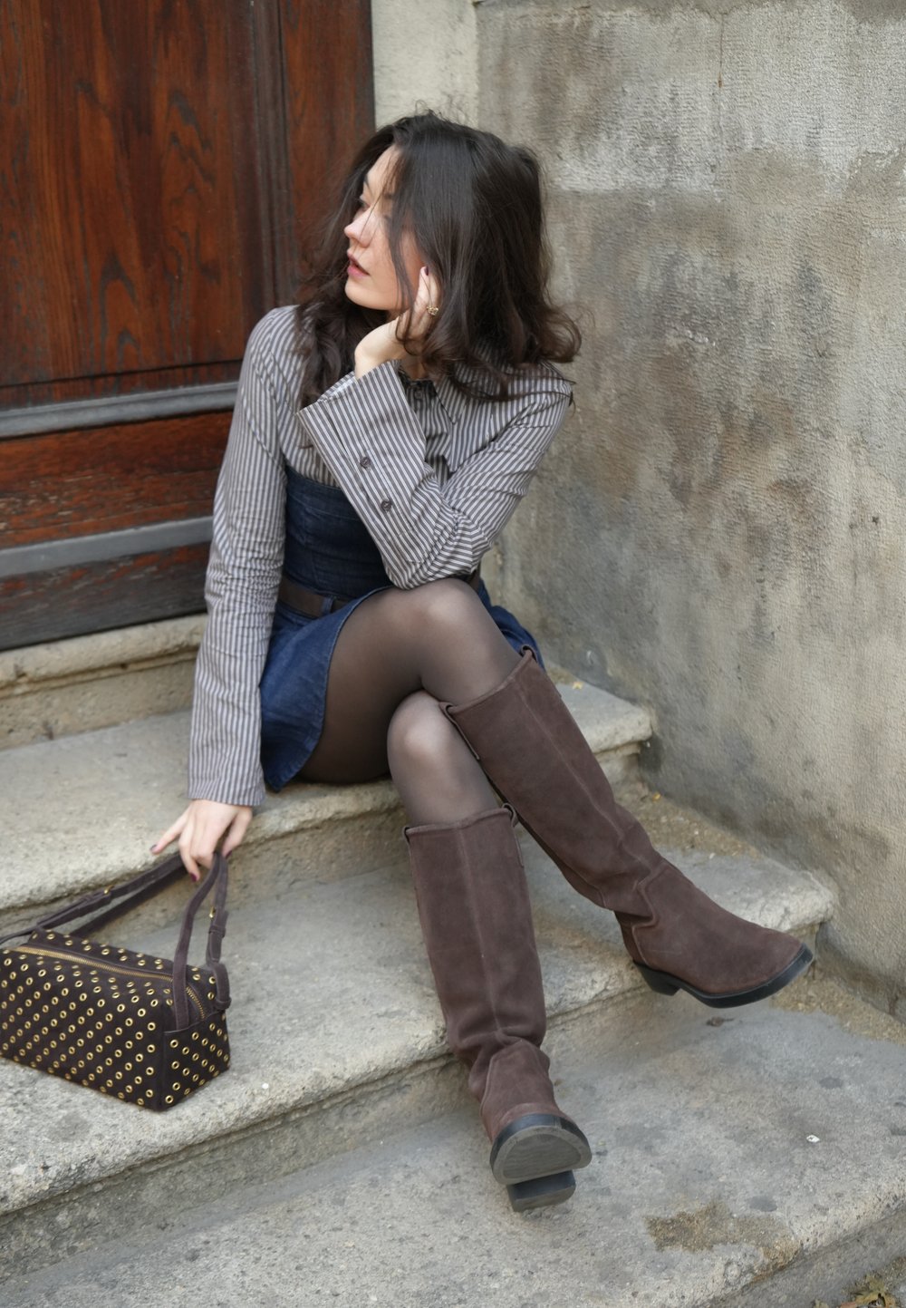 Bruine suède kniehoge laarzen, grijs gestreept shirt met lange mouwen, spijkerjurk en een donker patroon handtas op stenen trappen.
