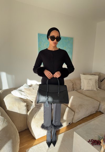 Cárdigan negro con puños de piel, pantalones grises con borde de piel, bolso negro con borla y zapatos negros puntiagudos en una habitación iluminada por el sol.