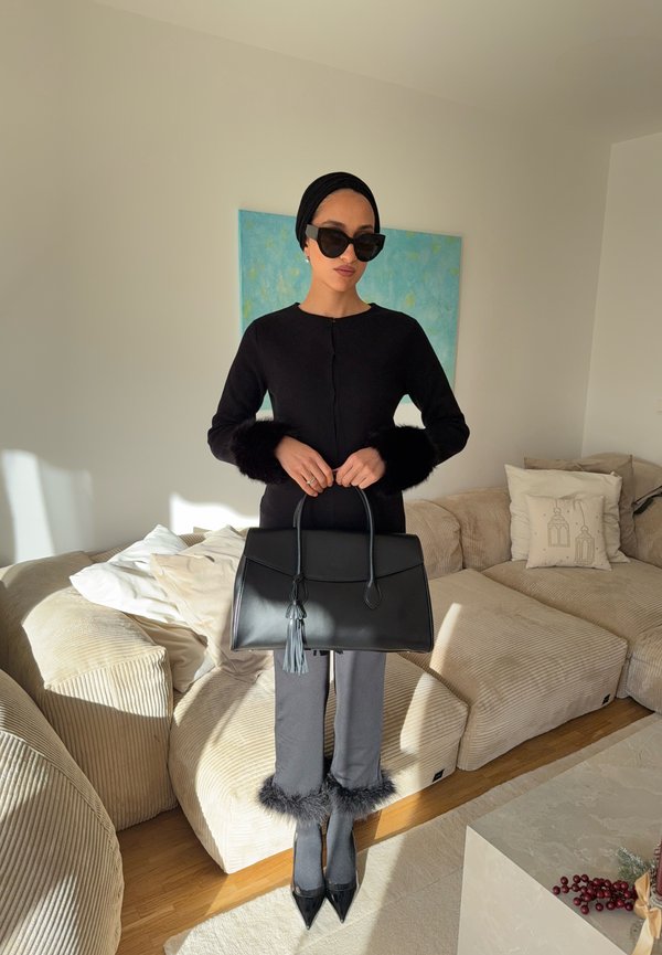Zwarte cardigan met bontmanchetten, grijze broek met bontafwerking, zwarte handtas met kwastje en zwarte puntige hakken in een zonovergoten kamer.