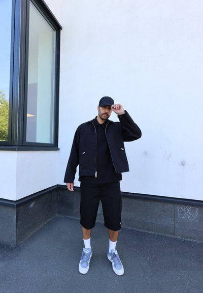 Chaqueta negra con cremallera sobre una camisa polo negra, pantalones cortos negros y zapatillas gris con suelas blancas. De pie frente a una pared blanca con ventanas.