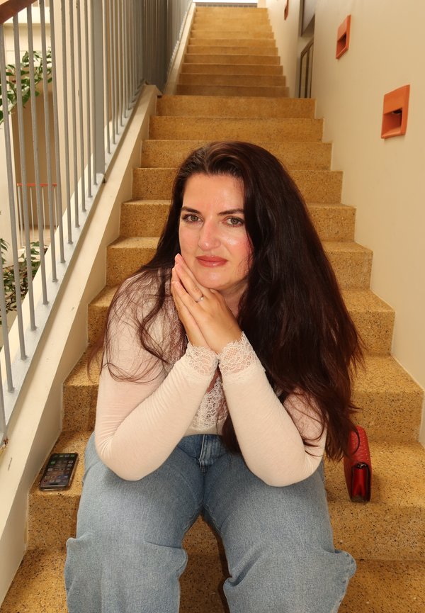 Femme aux longs cheveux foncés assise sur un escalier intérieur, reposant le menton sur ses mains jointes, avec un smartphone et un sac rouge à ses côtés.