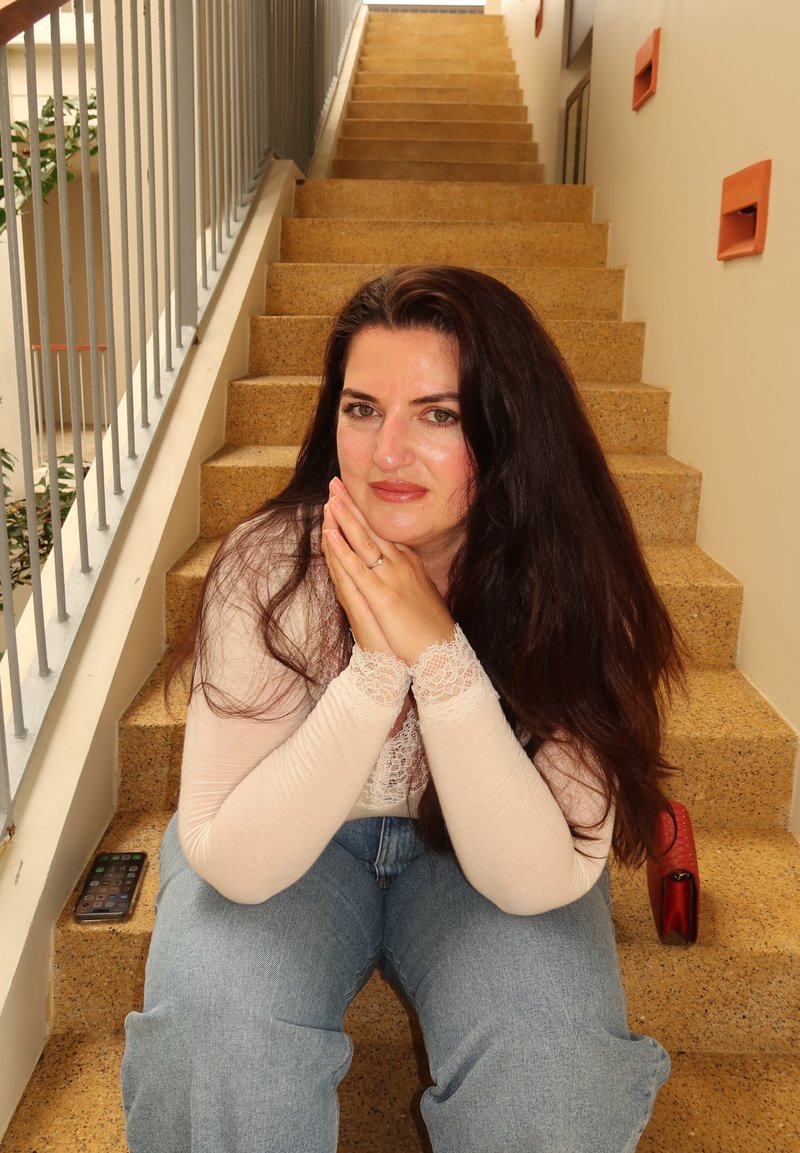 Femme aux longs cheveux foncés assise sur un escalier intérieur, reposant le menton sur ses mains jointes, avec un smartphone et un sac rouge à ses côtés.