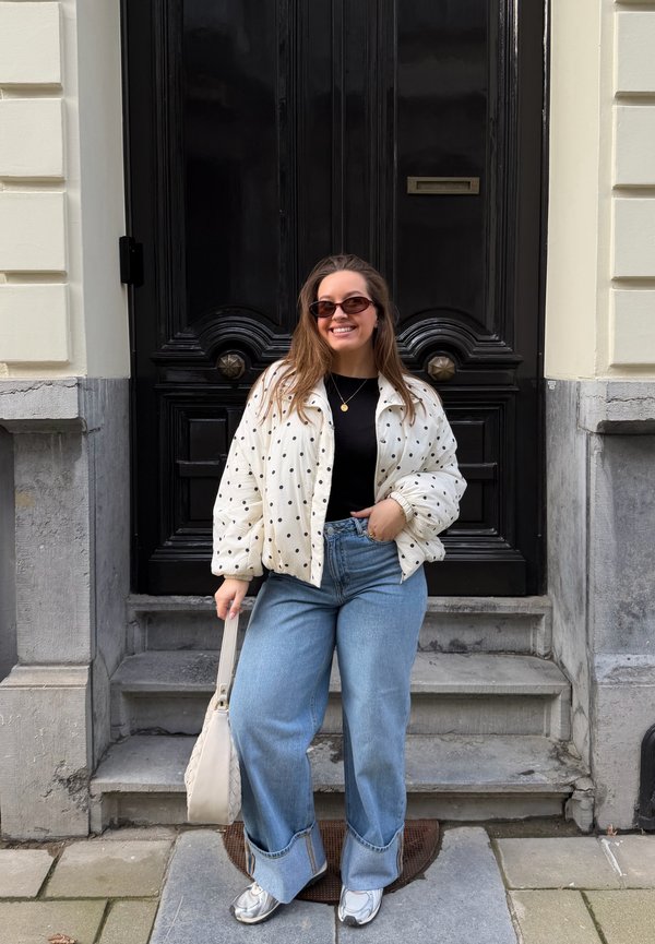 Naine naeratab päikeseprillidega, täpilise jakiga, musta topi, laiade säärega teksade, valgete tossude ja käekotiga, seisab kivist trepil musta ukse juures.