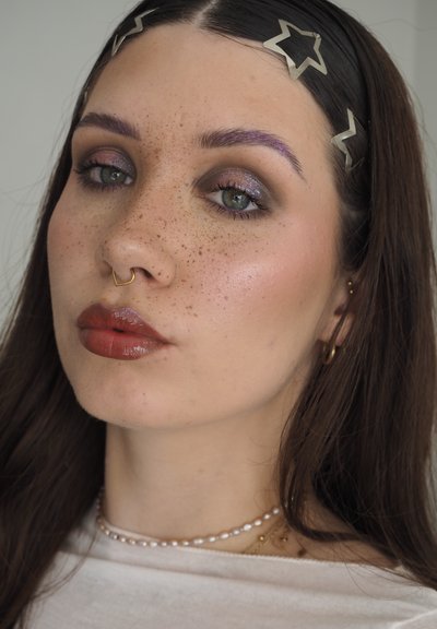 Joven mujer con pecas que lleva pinzas para el pelo en forma de estrella, anillo septum dorado, collar de perlas y pintalabios marrón brillante, con maquillaje de ojos brillante.