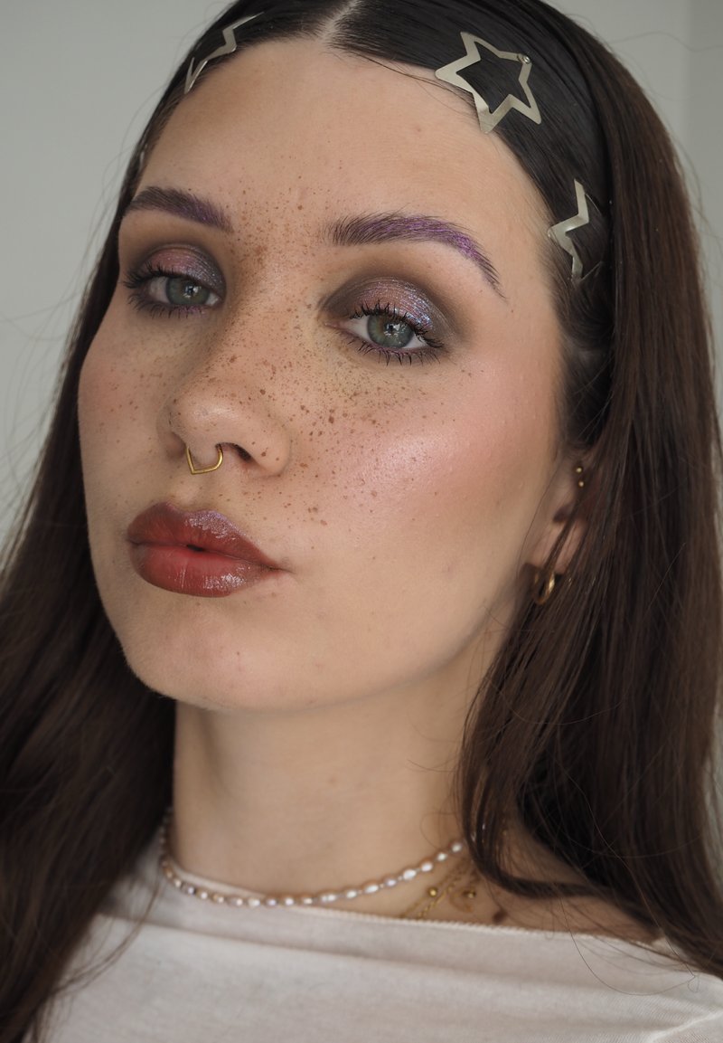 Jeune femme avec des taches de rousseur portant des barrettes en forme d’étoile, un anneau de septum en or, un collier de perles et un rouge à lèvres brun brillant, avec un maquillage des yeux scintillant.