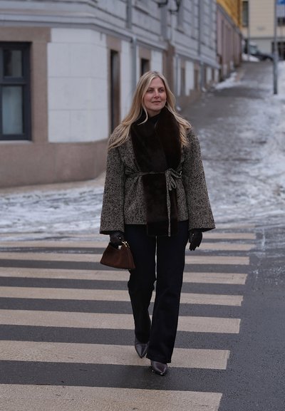 Mujer cruzando una calle nevada por un paso de cebra, llevando un abrigo con cinturón y cuello de piel, guantes negros, botas y un bolso marrón.