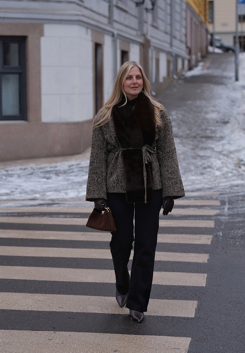 Γυναίκα που διασχίζει χιονισμένο δρόμο σε διάβαση πεζών, φορώντας παλτό με ζώνη και γούνινο γιακά, μαύρα γάντια, μπότες και κρατώντας καφέ τσάντα χειρός.