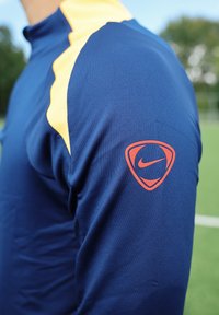 Person som bär en blå sportjacka med ett gult streck och en röd Nike-logotyp på den övre ärmen, står på en grön äng utomhus.