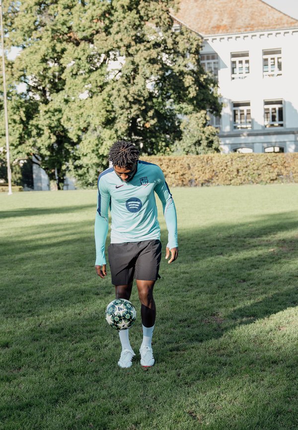 Un footballeur en maillot d’entraînement turquoise fait tenir en équilibre un ballon à motifs près de ses pieds sur un terrain herbeux, avec des arbres et un bâtiment derrière lui.