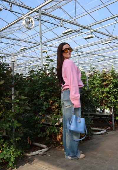 Mujer con suéter rosa y gafas de sol está en un invernadero sosteniendo un bolso azul entre plantas verdes altas.