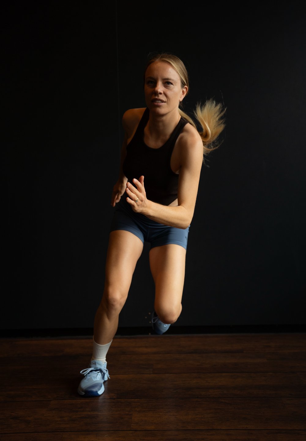 Atletische vrouw in een zwarte tanktop en blauwe shorts die aan het hardlopen is. Ze draagt lichtblauwe sneakers en witte sokken op een houten vloer.