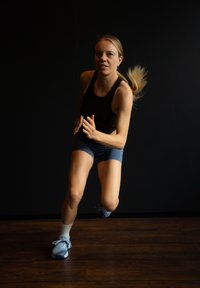 Atletische vrouw in een zwarte tanktop en blauwe shorts die aan het hardlopen is. Ze draagt lichtblauwe sneakers en witte sokken op een houten vloer.