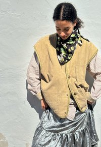 Beige kordflorsväst med knappar framtill, lagrad över en rutig skjorta. Svart scarf med blad- och kaninmönster. Glänsande silverkjol.