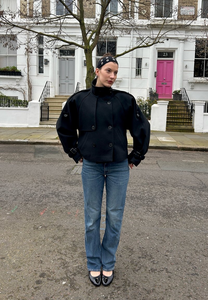 Frau mit kurzen Haaren, die eine schwarze Jacke, blaue Jeans und schwarze Schuhe trägt, steht auf einer Straße vor weißen Stadthäusern mit grauen und rosa Türen.