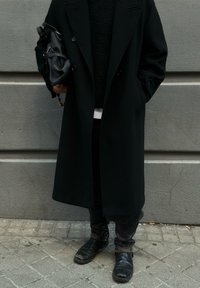 Abrigo largo de lana negro con diseño cruzado, combinado con un suéter negro texturizado, pantalones negros y botines negros.