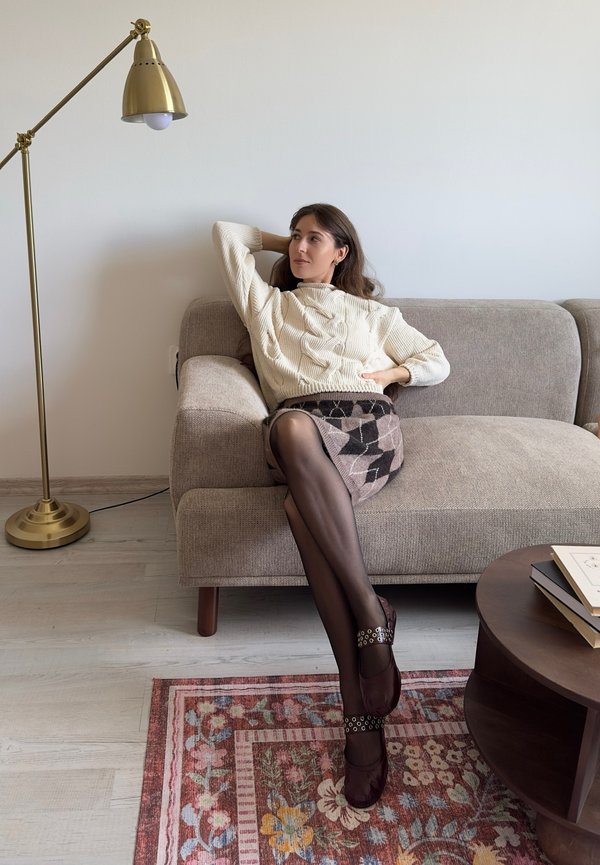 Pull crème tricoté, jupe brune à motifs, collants noirs et chaussures bordeaux avec des accents circulaires. Assise sur un canapé en tissu gris clair.