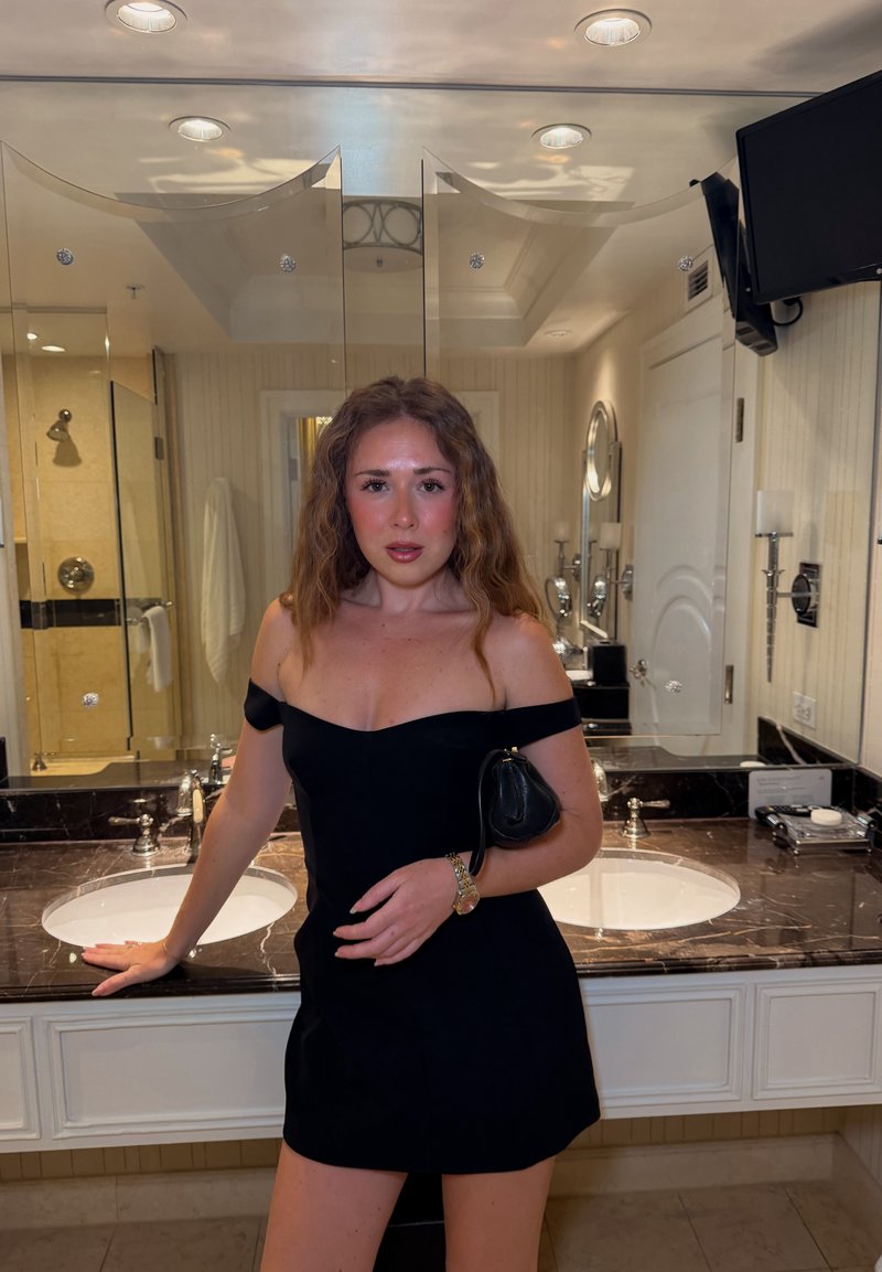 Femme en robe noire à épaules dénudées, debout dans une salle de bains avec un plan de travail en marbre, deux éviers, des miroirs et une télévision murale.