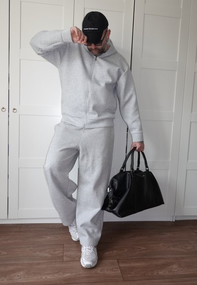 Hombre con chándal gris y zapatillas blancas sosteniendo una bolsa negra grande, ajustándose una gorra negra, de pie sobre un suelo de madera frente a armarios blancos.