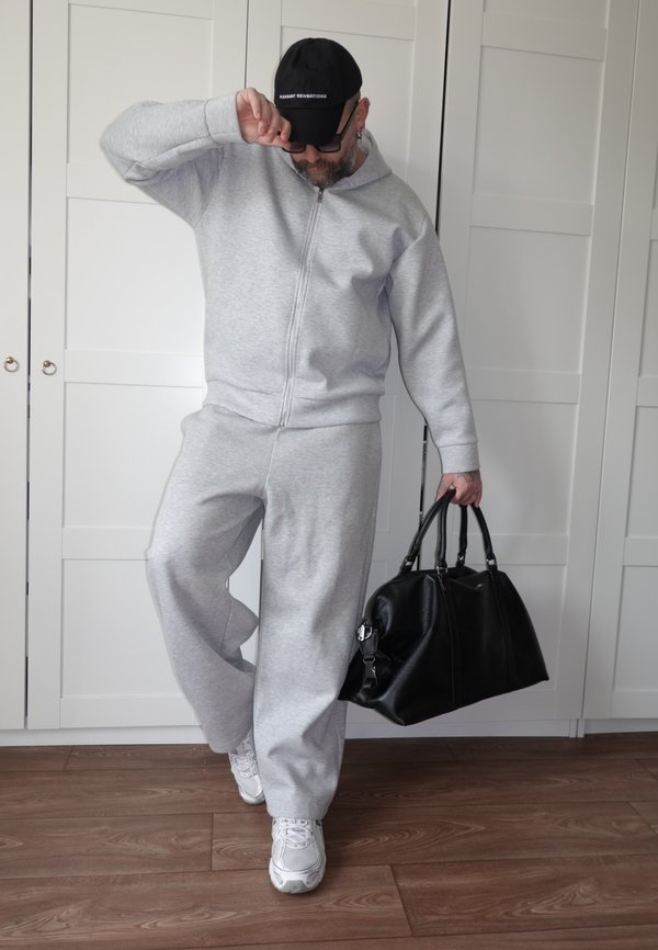 Muškarac u sivom trenirkom i bijelim tenisicama drži veliku crnu torbu, podešava crnu kapu, stoji na drvenom podu ispred bijelih ormara.