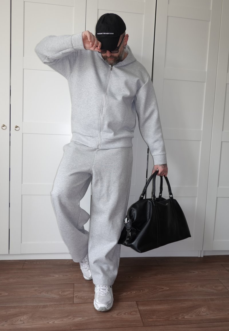 Muž v sivom teplákovom odeve a bielych teniskách drží veľkú čiernu tašku, upravuje si čiernu šiltovku, stojí na drevenej podlahe pred bielymi skrinkami.