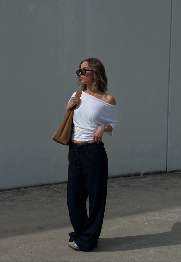 Vrouw met kort golvend haar draagt een witte off-shoulder top, donkere wijde broek, zonnebril en draagt een bruine tote bag, staand op beton bij een grijze muur.