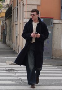 Uma pessoa com um longo casaco escuro azul-marinho sobre uma camisa creme, calças de ganga de perna larga e sapatos castanhos, segurando um smartphone enquanto caminha.
