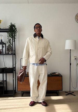 Hombre con chaqueta y pantalones color crema, sosteniendo una bolsa marrón en una sala de estar minimalista con una estantería negra y una lámpara de pie.