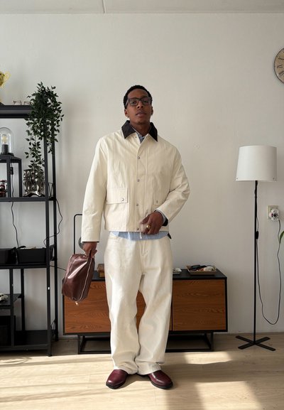 Hombre con chaqueta y pantalones color crema, sosteniendo una bolsa marrón en una sala de estar minimalista con una estantería negra y una lámpara de pie.